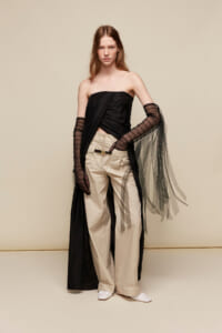 Female model in a black strapless top, beige wide-leg pants, and a sheer black cape with mesh gloves against a neutral backdrop.