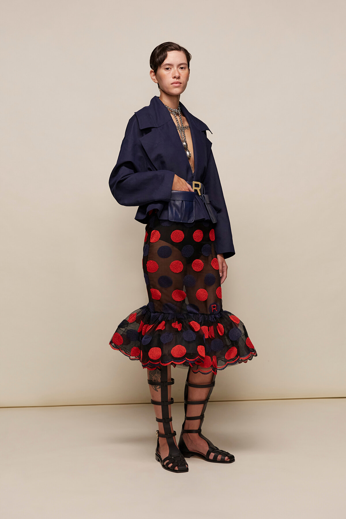 Model in a navy oversized jacket with a sheer polka-dot skirt (red and navy dots), ruffled hem, belt bag, and strappy sandals.