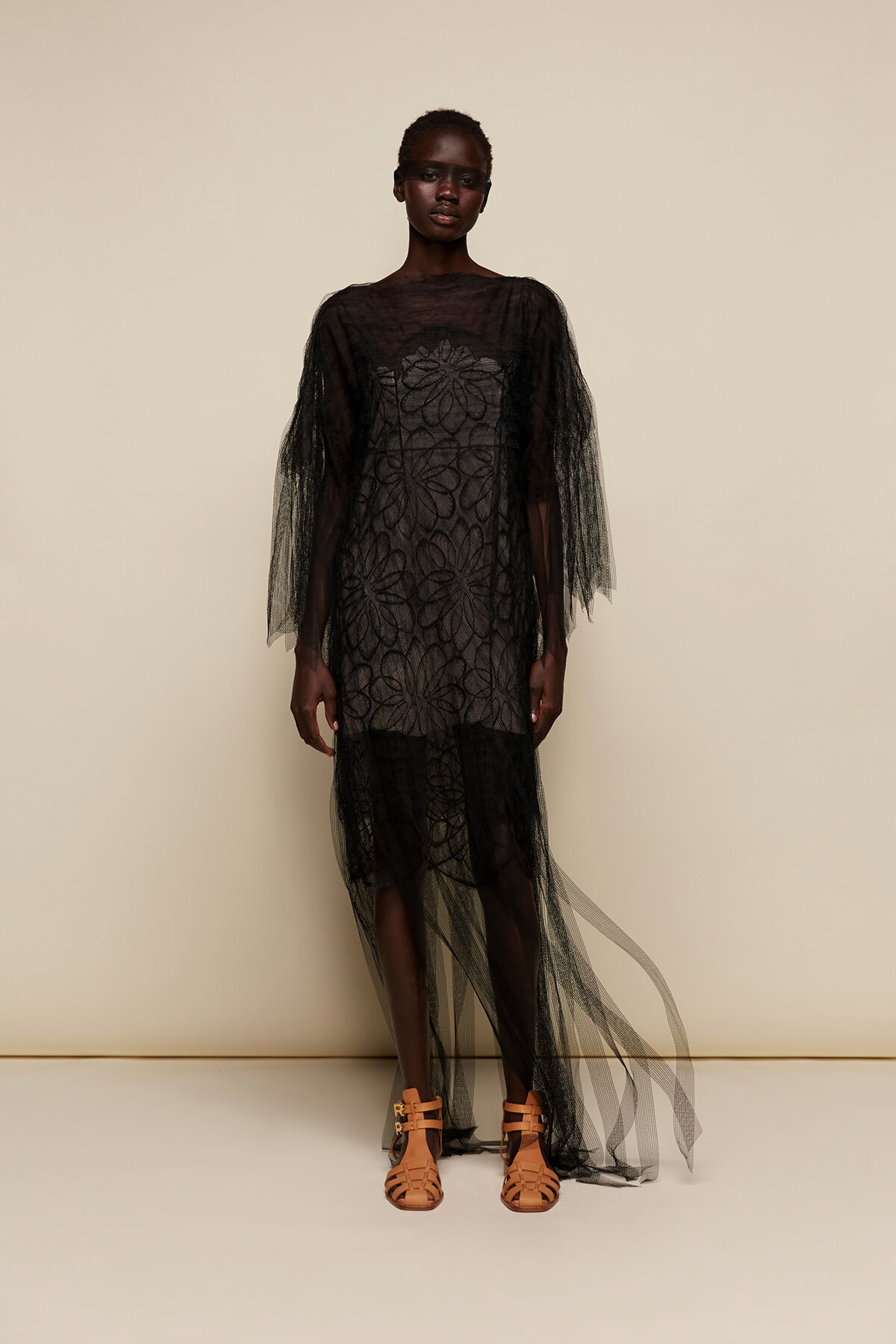 Models wearing a sheer black lace gown with layered cape and tan gladiator sandals, standing against a neutral backdrop.