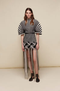 Model wearing a black-and-white striped top with large cape-like sleeves, cinched at the waist, paired with a sheer plaid skirt and black sandals.