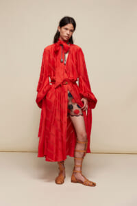 Woman wearing a red satin dress with an oversized bow scarf, belt, and tan gladiator sandals against a neutral backdrop.