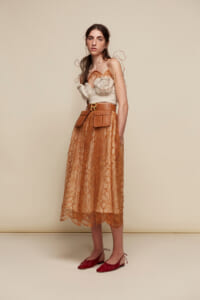 Model wearing a beige bustier top with an orange lace midi skirt, tan belt with pockets, and red flat shoes against a neutral backdrop.