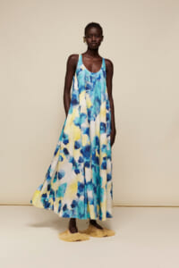 Model wearing a sleeveless blue-yellow-white tie-dye maxi dress with fringe sandals in a beige studio setting