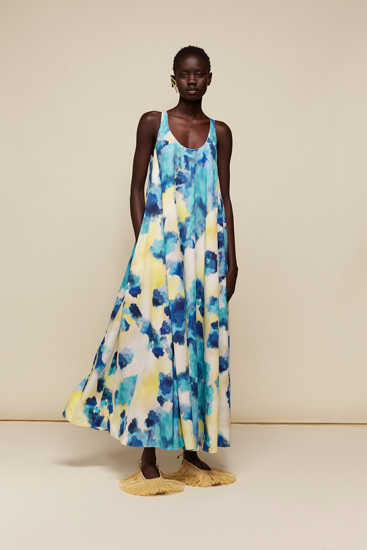 Model wearing a sleeveless blue-yellow-white tie-dye maxi dress with fringe sandals in a beige studio setting