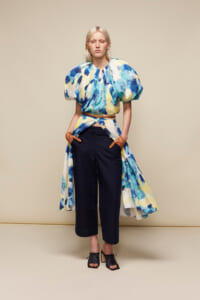 Model in a blue-yellow tie-dye top with puff sleeves, belted at the waist, navy cropped trousers, and black open-toe sandals.