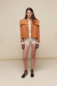 Model wearing an orange textured cropped jacket over a white blouse and cream shorts, with black strappy sandals against a beige backdrop.