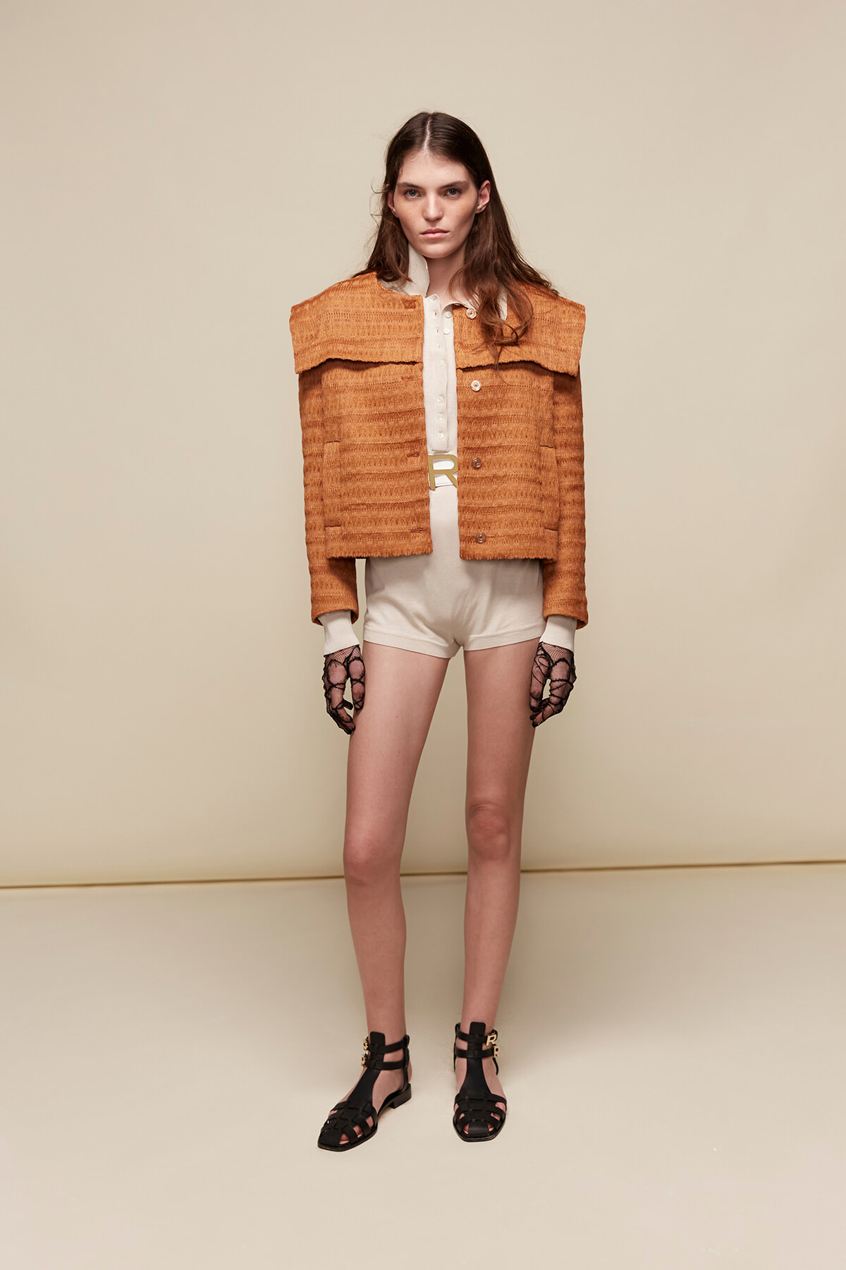 Model wearing an orange textured cropped jacket over a white blouse and cream shorts, with black strappy sandals against a beige backdrop.