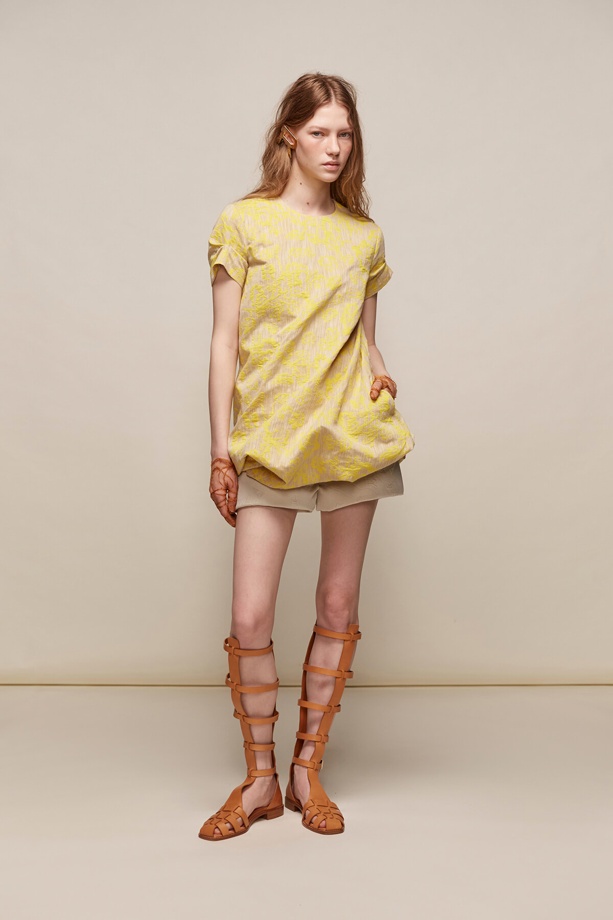 Model wearing a pale yellow floral tunic over beige shorts with tan gladiator sandals.