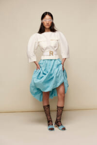 Model wearing a white top with oversized sleeves and a belted waist, paired with a voluminous light-blue skirt, lace tights, and blue heeled sandals; eyes covered with a blindfold for a fashion editorial look.
