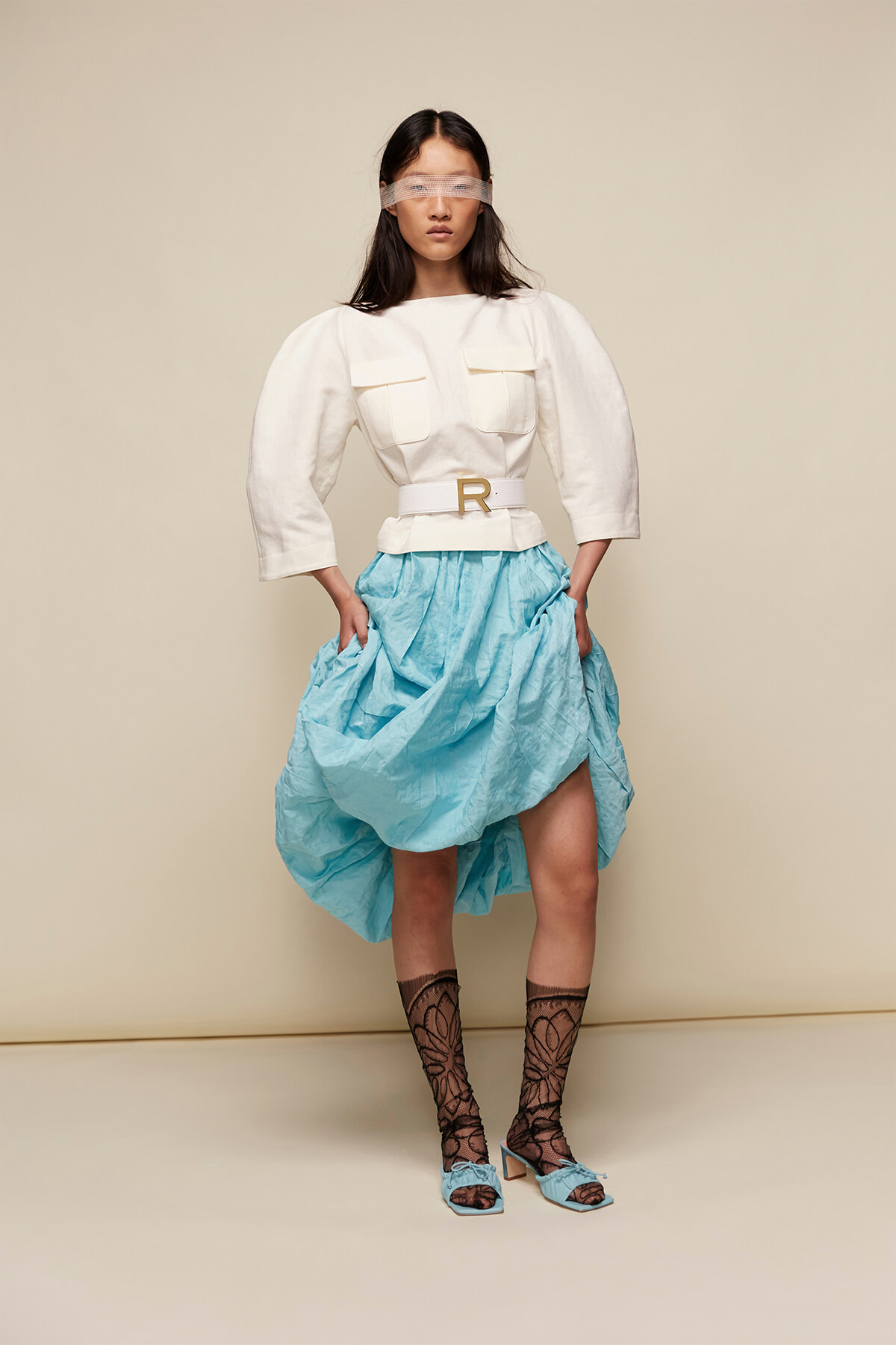 Model wearing a white top with oversized sleeves and a belted waist, paired with a voluminous light-blue skirt, lace tights, and blue heeled sandals; eyes covered with a blindfold for a fashion editorial look.