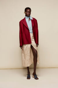 Model wearing a burgundy oversized blazer over a light-blue polo, paired with a crinkled beige wrap skirt and heeled sandals against a neutral beige backdrop.