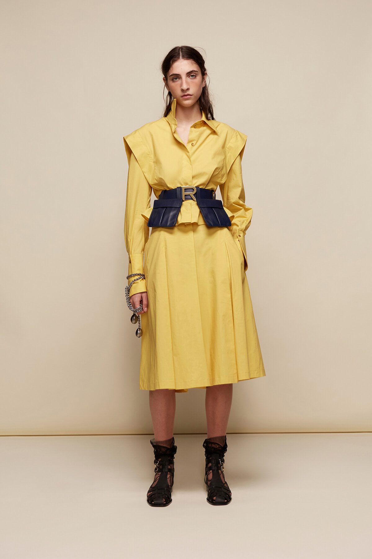 Model wearing a bright yellow midi dress with oversized shoulders and a wide navy belt, standing against a beige backdrop.