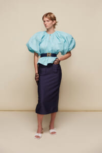 Model wearing a light blue puff-sleeve blouse with a navy belt and navy pencil skirt, standing against a beige backdrop.