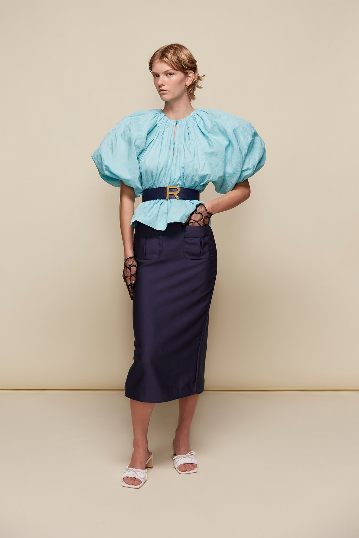 Model wearing a light blue puff-sleeve blouse with a navy belt and navy pencil skirt, standing against a beige backdrop.