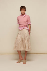 Model wearing a pink knit top with ruffled pink blouse peeking at the hem, beige crinkled midi skirt, and open-toe heeled sandals against a neutral backdrop.