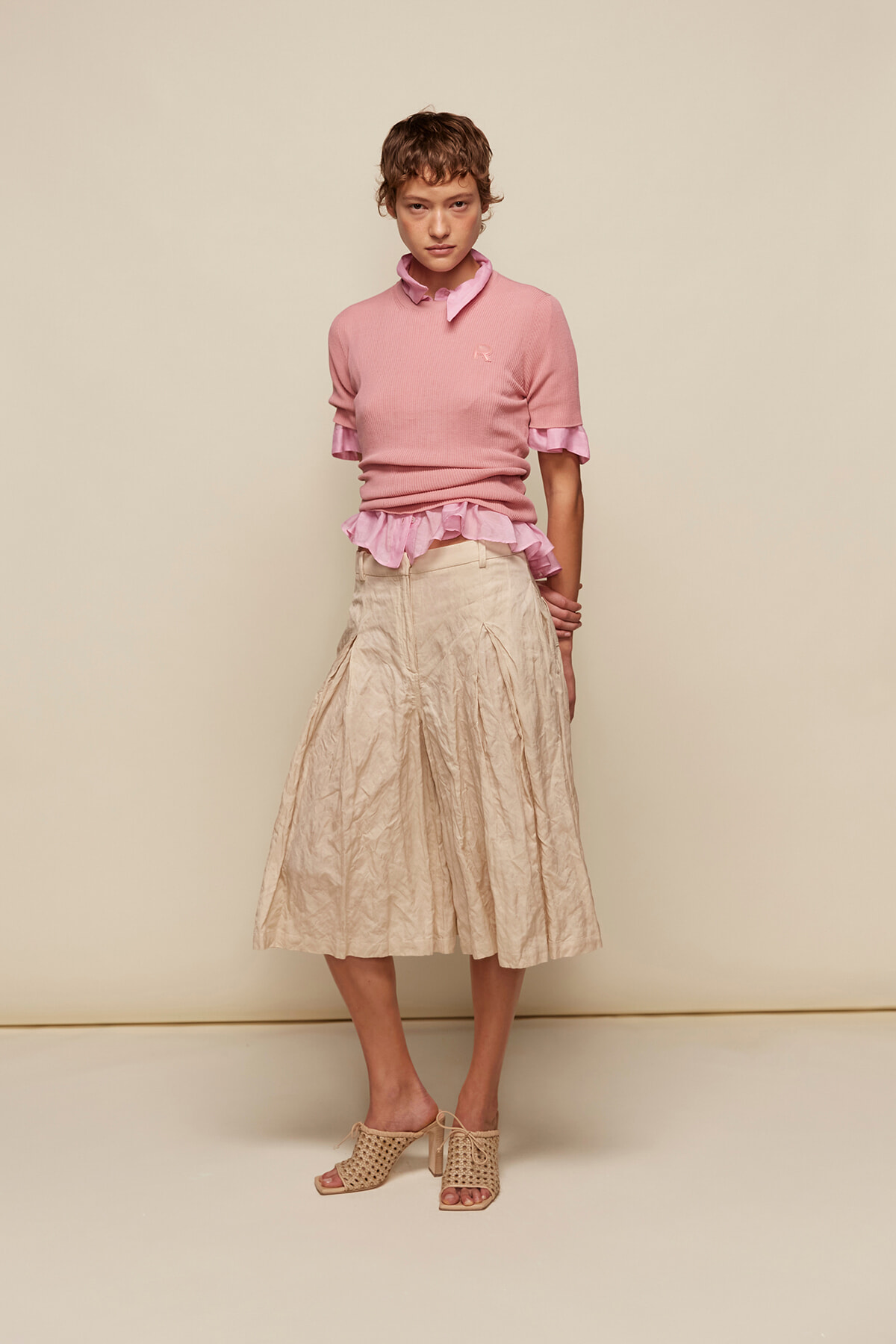 Model wearing a pink knit top with ruffled pink blouse peeking at the hem, beige crinkled midi skirt, and open-toe heeled sandals against a neutral backdrop.