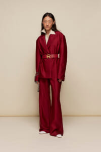 Person wearing a burgundy blazer and wide-leg trousers with a matching belt and cream shirt, posing against a beige backdrop.