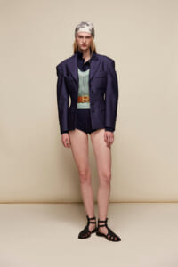 Model wearing an oversized navy blazer over a mint knit top with a tan belt, high-cut navy shorts, and black strappy sandals, plus a patterned headscarf, standing against a beige backdrop.