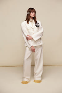 Model in an oversized white suit with a beige backdrop, wearing a blindfold and one arm crossed over the other, posing for a fashion shoot.
