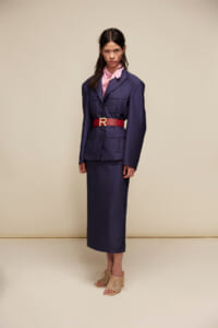 Model wearing a navy utility-style suit with a pink blouse, red belt with a gold buckle, and woven beige sandals against a beige backdrop.