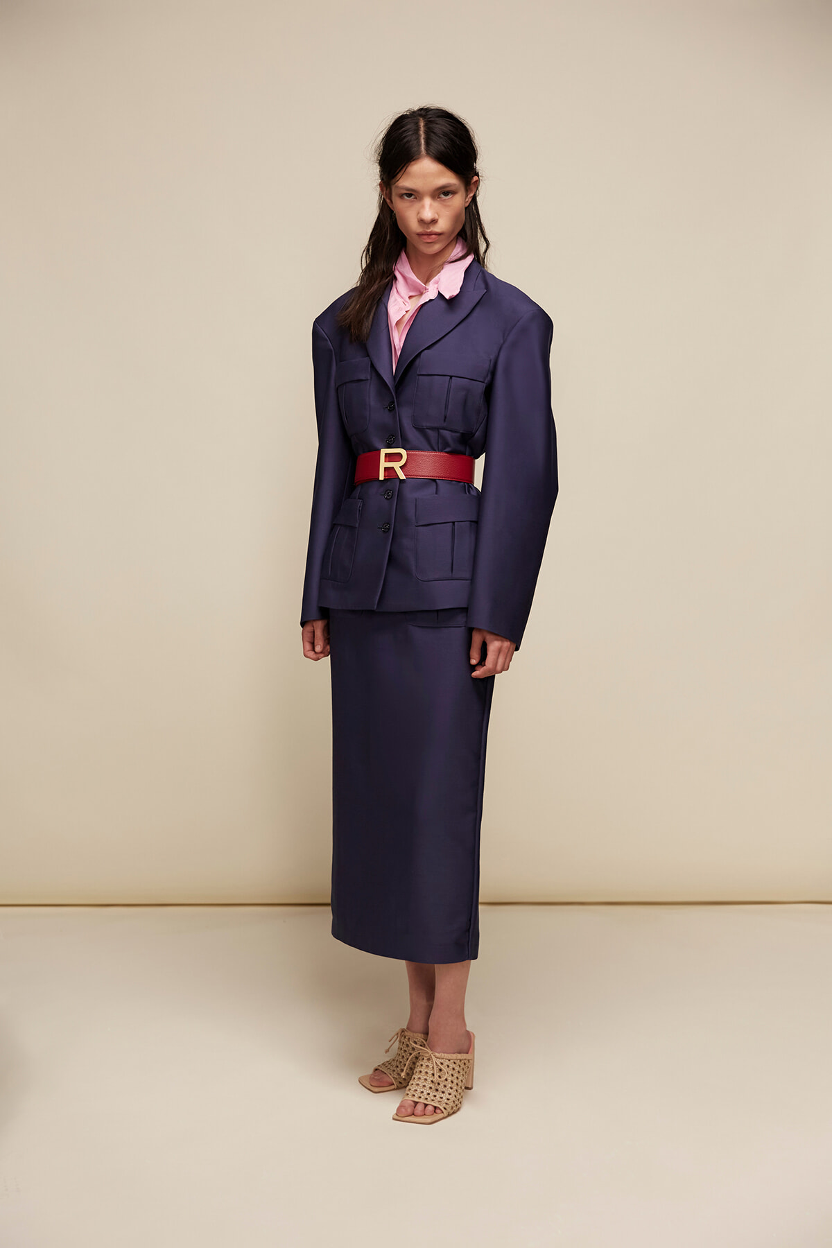 Model wearing a navy utility-style suit with a pink blouse, red belt with a gold buckle, and woven beige sandals against a beige backdrop.