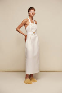 Model in a white halter-neck maxi dress with a belt, standing in beige studio with fringed yellow sandals and a sheer eye cover.