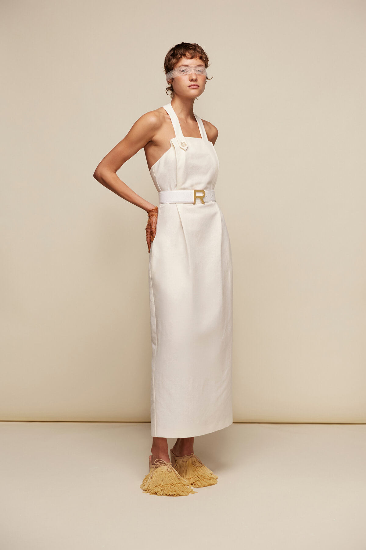 Model in a white halter-neck maxi dress with a belt, standing in beige studio with fringed yellow sandals and a sheer eye cover.