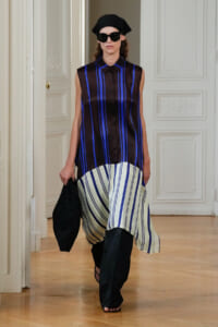 Model walking the runway in a sleeveless black-and-blue striped tunic with a white striped hem, black headscarf, oversized sunglasses, and a black clutch.