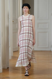 Model walking in a sleeveless white knit maxi dress with pink and burgundy plaid grid, paired with a black headscarf and strappy sandals on a fashion runway.