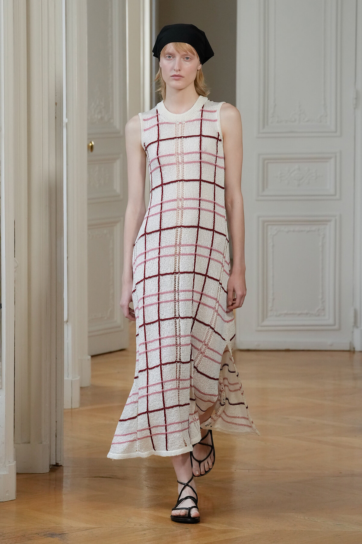 Model walking in a sleeveless white knit maxi dress with pink and burgundy plaid grid, paired with a black headscarf and strappy sandals on a fashion runway.