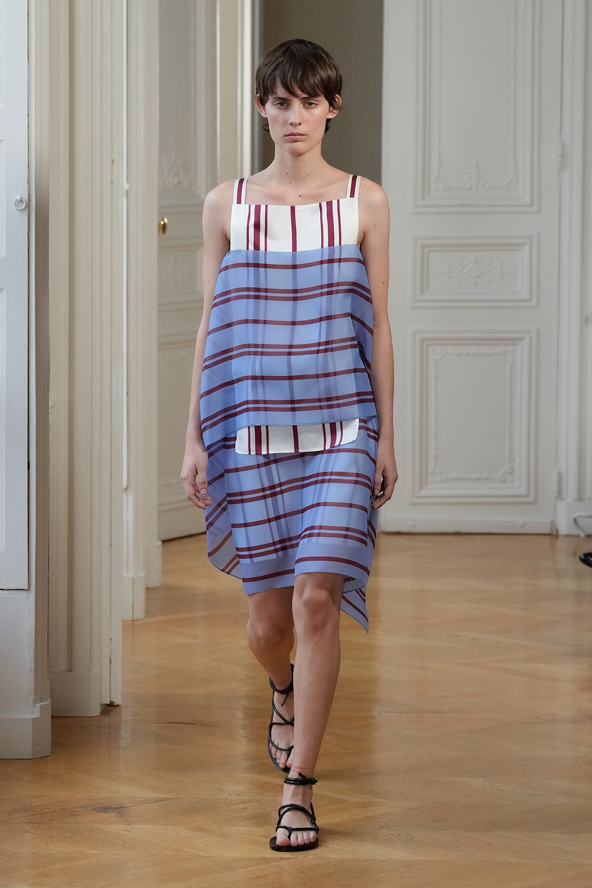 Model walking a fashion runway wearing a loose, layered blue and white striped dress with red accents; white top with vertical stripes peeks from underneath. She pairs with black strappy sandals in a sunlit, ornate room.
