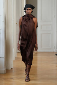 Female model walks a runway in a dark brown satin draped dress, with a black headscarf and strappy sandals in a bright, ornate room.