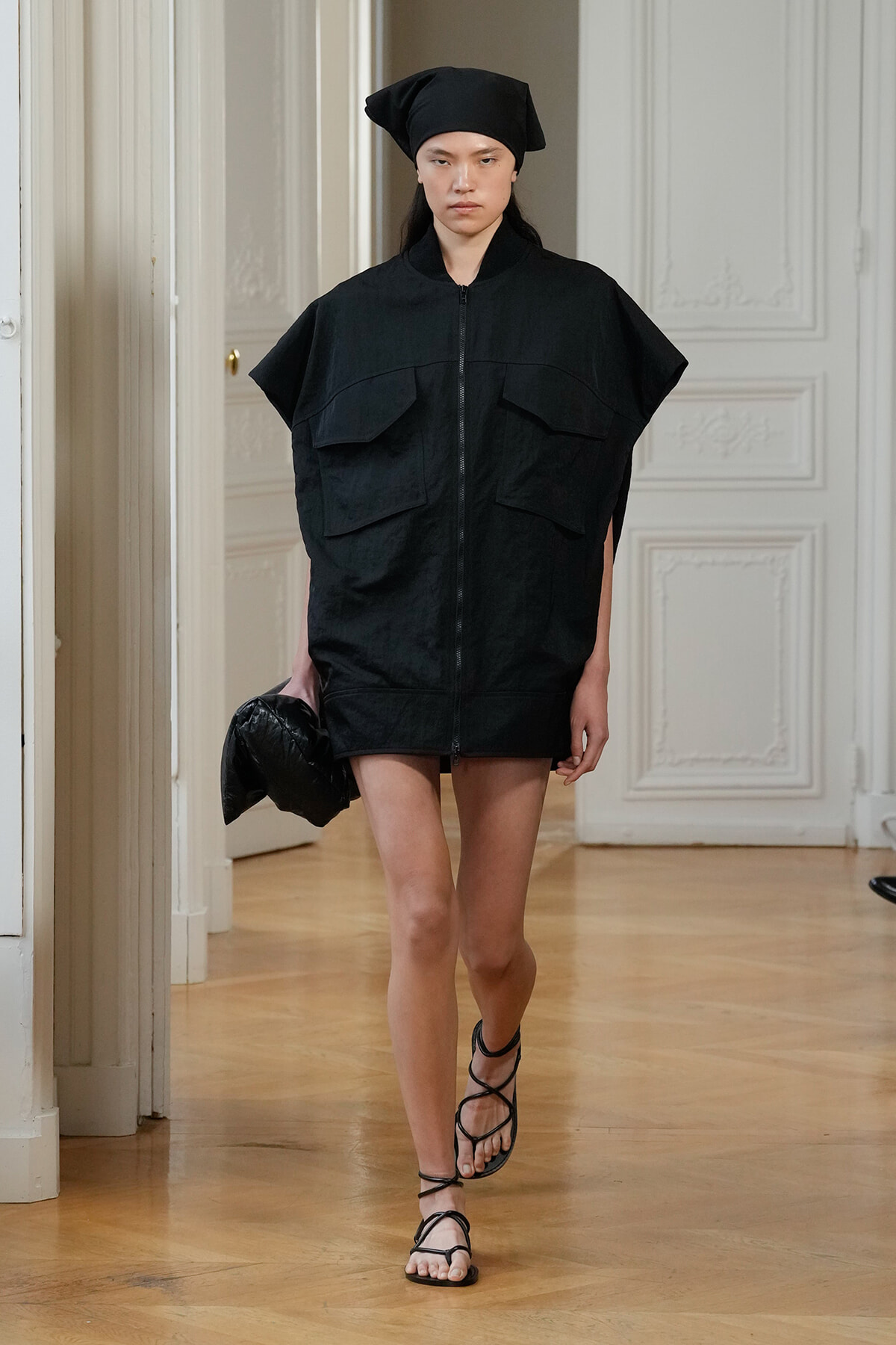 Female model walks a runway in an oversized black zip-front jacket with large front pockets, black head wrap, and strappy sandals, carrying a black bag as she models.",
