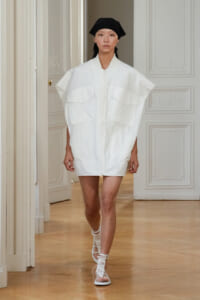 Model walks in a white oversized zip-front dress with large front pockets, black headscarf, and white crochet-strap sandals in a bright, ornate room.