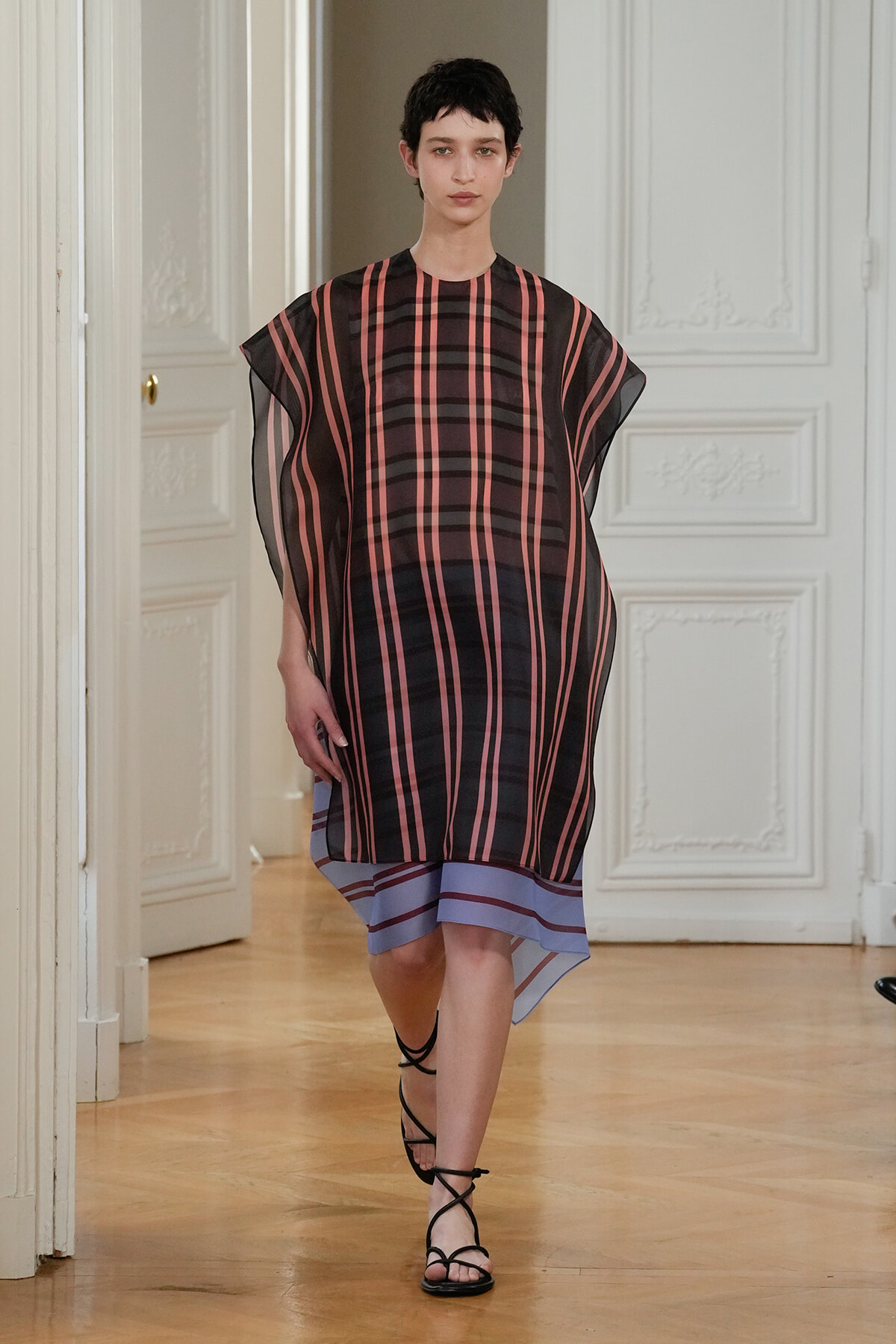 Model walks down a runway in a sheer, oversized striped tunic-dress with pink and black vertical stripes, blue layered hem, and black strappy sandals in a white-paneled room.