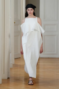 Model wearing a white off-shoulder dress with a large ruffle, black headscarf, and strappy flat sandals walking in a formal room.