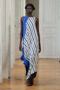 Model in a blue and white striped draped dress with a black head wrap walking the runway in a white, ornate room.