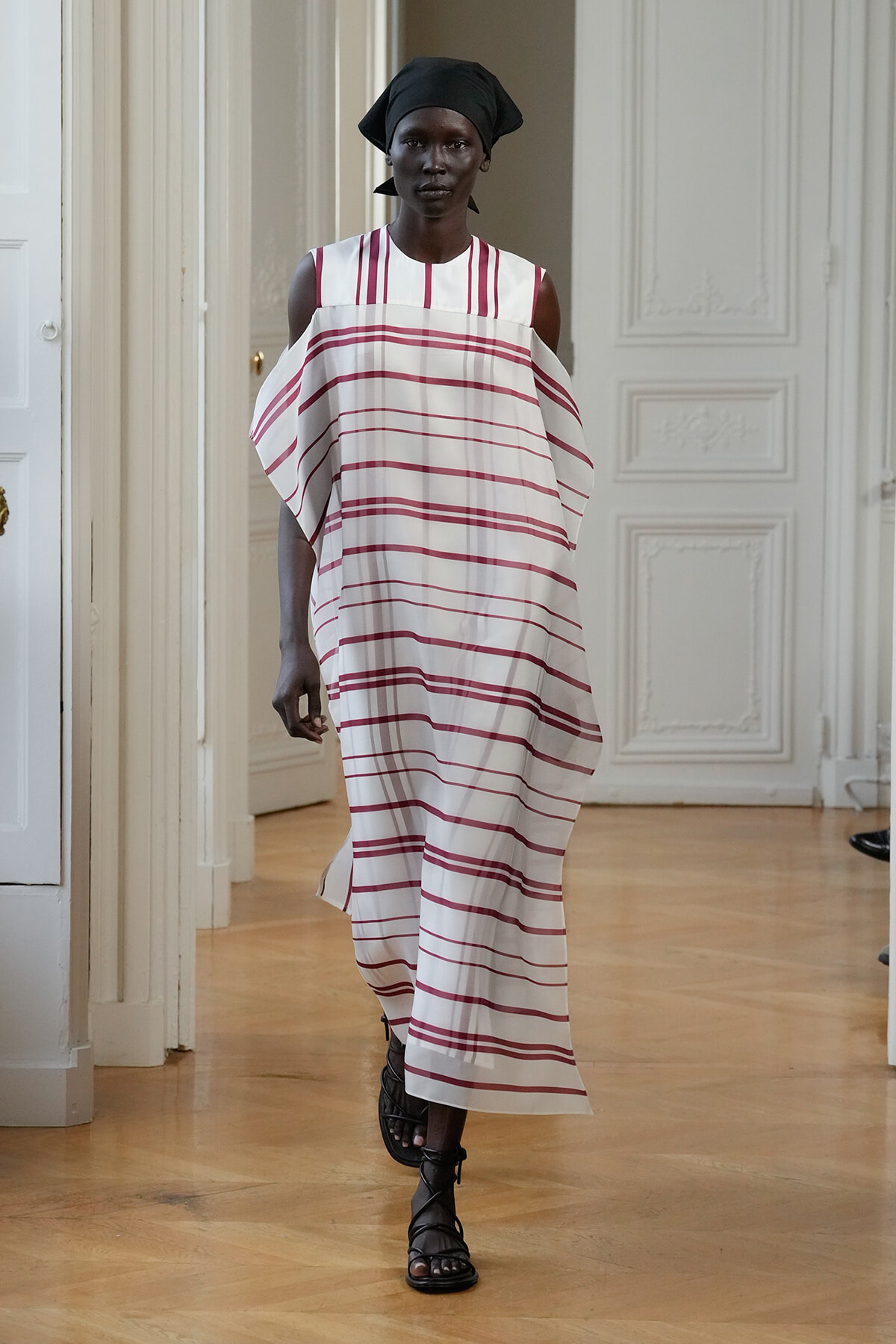 Model wearing a white dress with red stripes and a black headscarf walking down a light-painted hallway runway style.