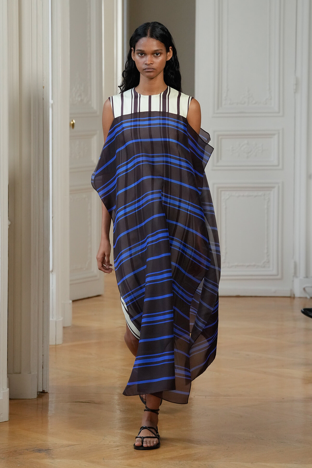 Model walking in a neutral room wearing a loose, draped brown and blue striped poncho-style dress over a sleeveless striped top, paired with black flat sandals.