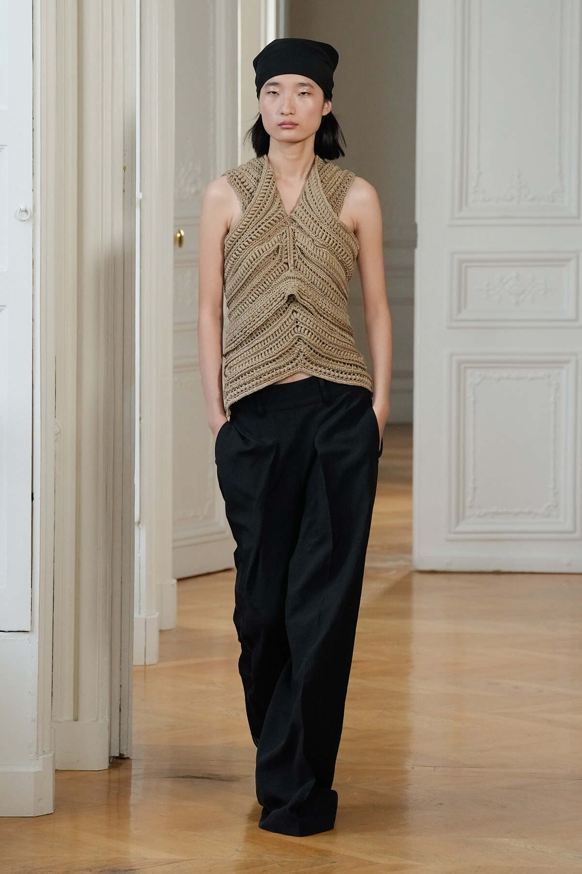 Model wearing a beige crocheted halter top and black wide-leg pants stands in a doorway with ornate white walls.