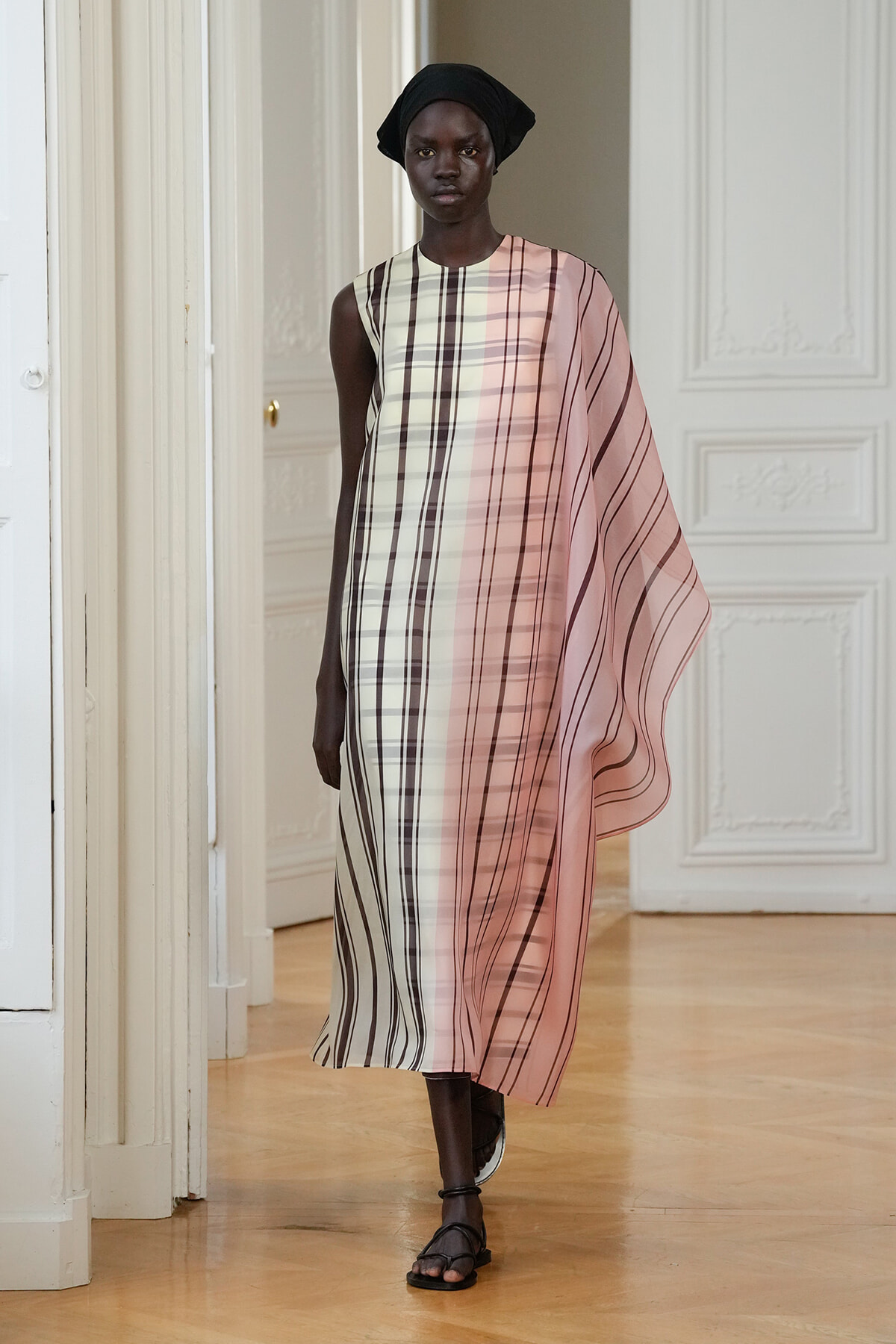 Model walking in a white-paneled room wearing a flowing pink plaid dress with a draped cape-like panel and a black headwrap, black sandals.