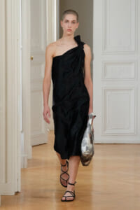 Model wearing a black one-shoulder draped dress, strappy sandals, and a patterned tote, walking through a bright, ornate room.