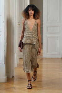 Model walking in a beige crochet halter dress with a layered top and cropped wide-leg skirt, carrying a brown clutch and wearing black strappy sandals.