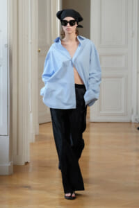Model walking in a light blue oversized shirt, black wide-leg pants, black headscarf and sunglasses in a bright room with wood flooring. (informative)