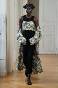 Model wearing a black knit dress with a floral bustier and a long floral-patterned coat, head wrapped in black fabric, standing in a bright room.
