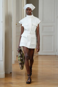 Model wearing a white utility dress with pockets and belt, plus a white headwrap, walking in a bright room.