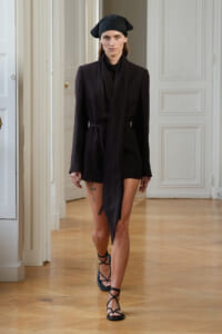 Model wearing an all-black blazer-style mini dress with a belt and a black headwrap, walking in a classical white interior.