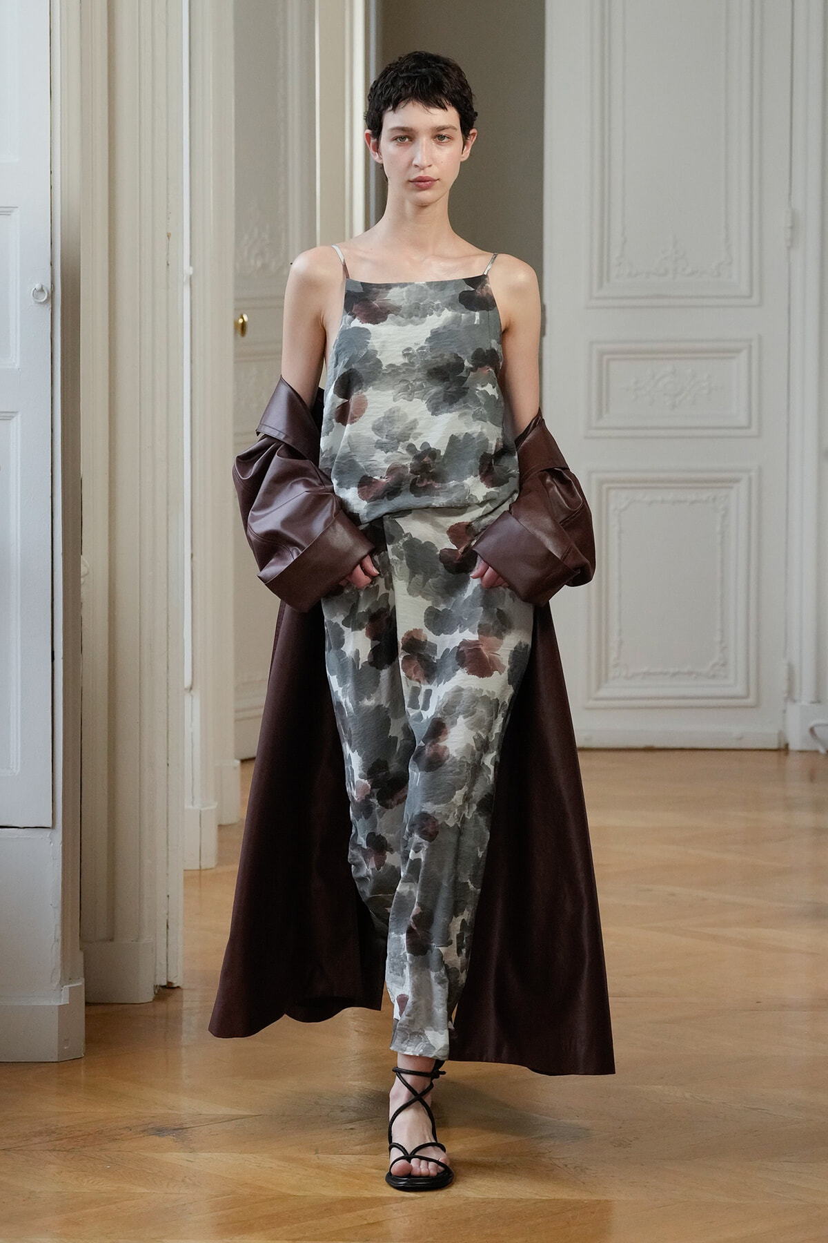 Model wears a gray floral sleeveless jumpsuit with a dark brown long coat draped over her arms, standing on a wooden floor indoors.