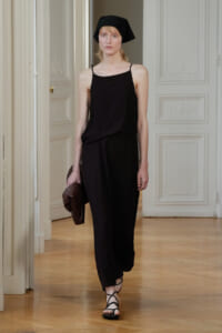 Model walking a runway in a black spaghetti-strap jumpsuit, black headscarf, strappy sandals, and holding a brown jacket in a pale ornate hallway