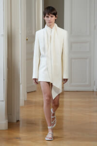 Model wearing an all-white tailored blazer with a draped scarf walking down a wooden-floor runway.